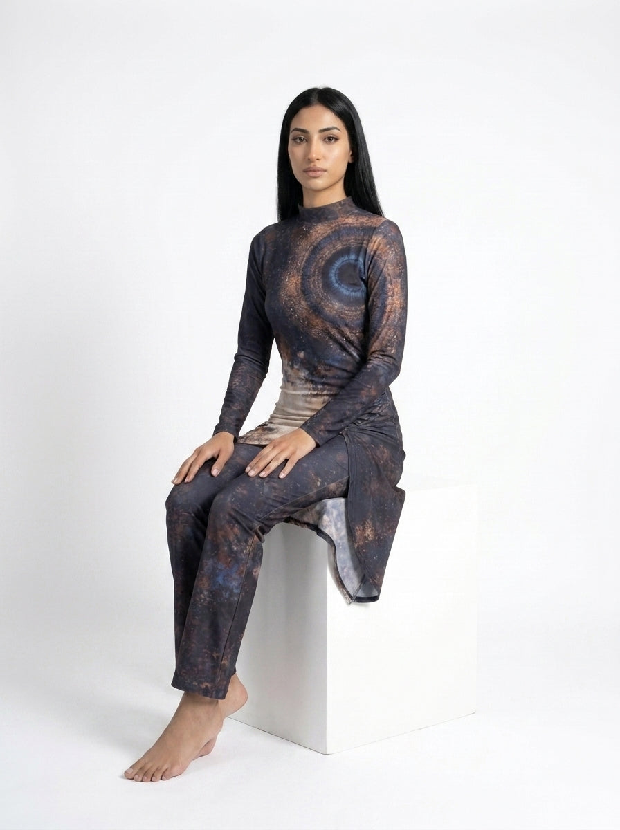 A woman sits barefoot on a white block against a white studio background, wearing a long-sleeve mock-neck tunic and matching pants set in a dark abstract print with brown, blue, and gray tones.