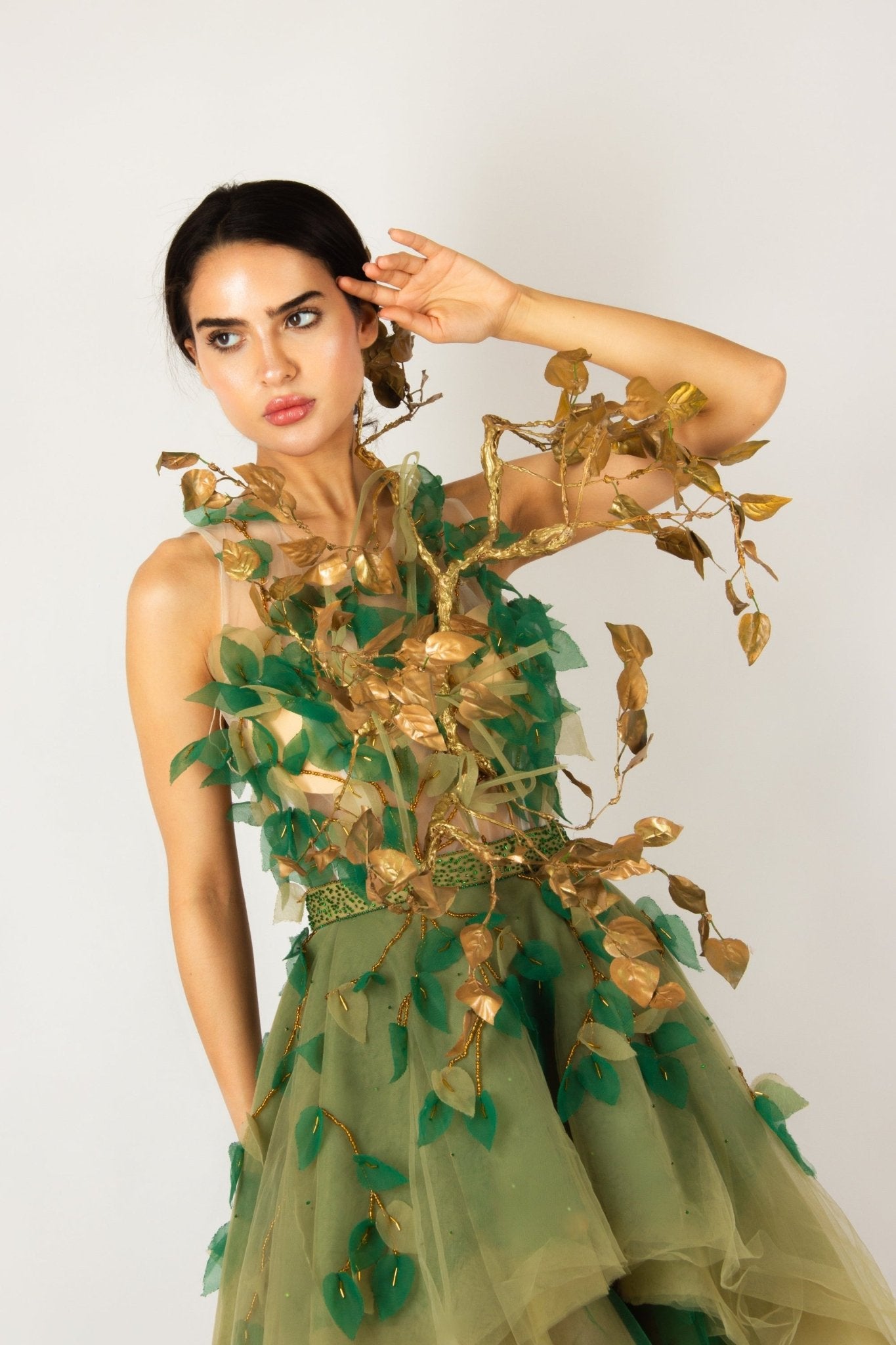 Enchanting nature-inspired gown by MNour Design, featuring a flowing olive-green tulle skirt with a high-low cut and an intricately designed bodice adorned with gold and green leaf appliqués. A breathtaking fusion of fashion and artistry. Discover this exclusive design at www.mnourdesign.com