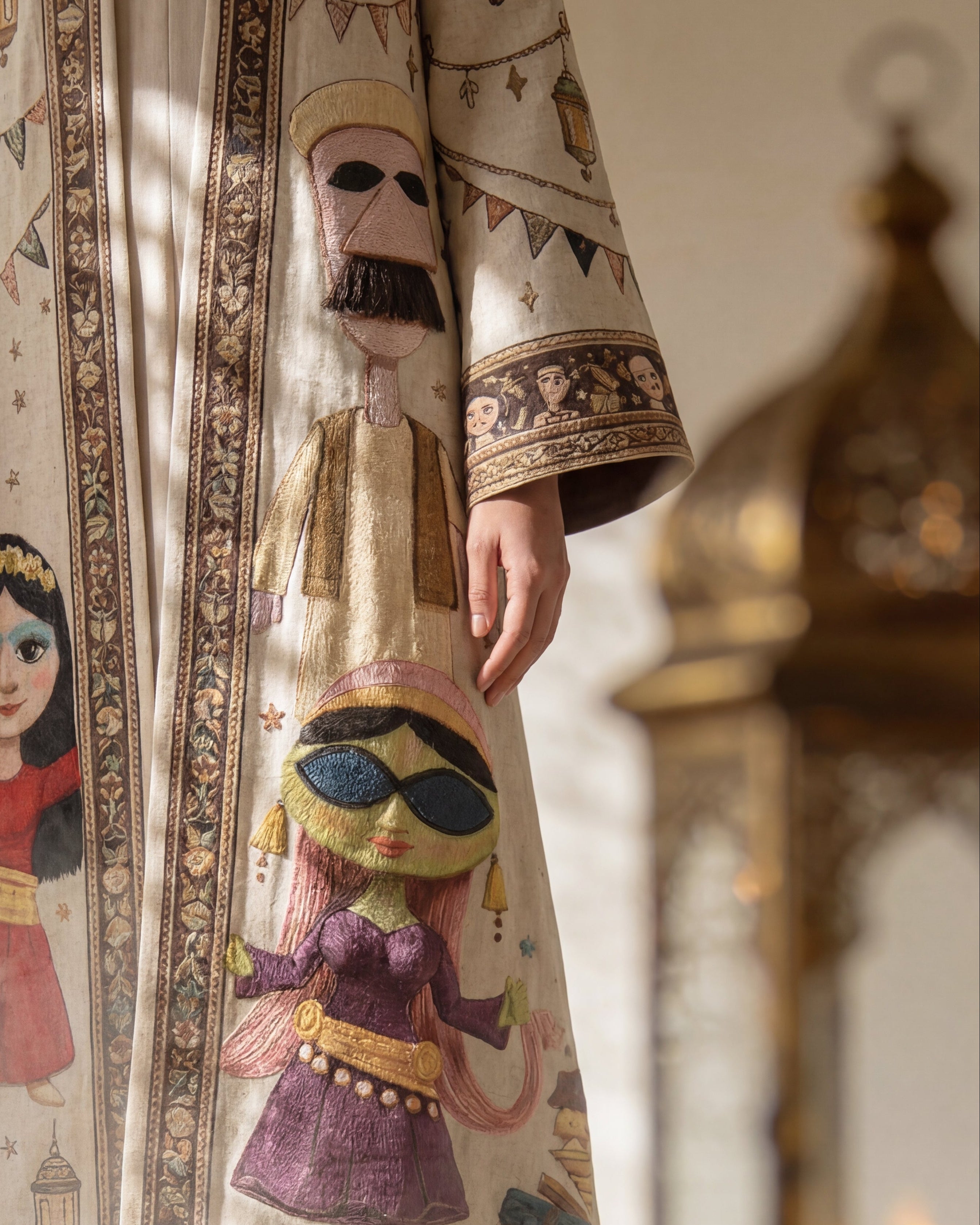 Traditional garment with intricate designs of people and lanterns, held by a person, with a candlelit lantern in the background.