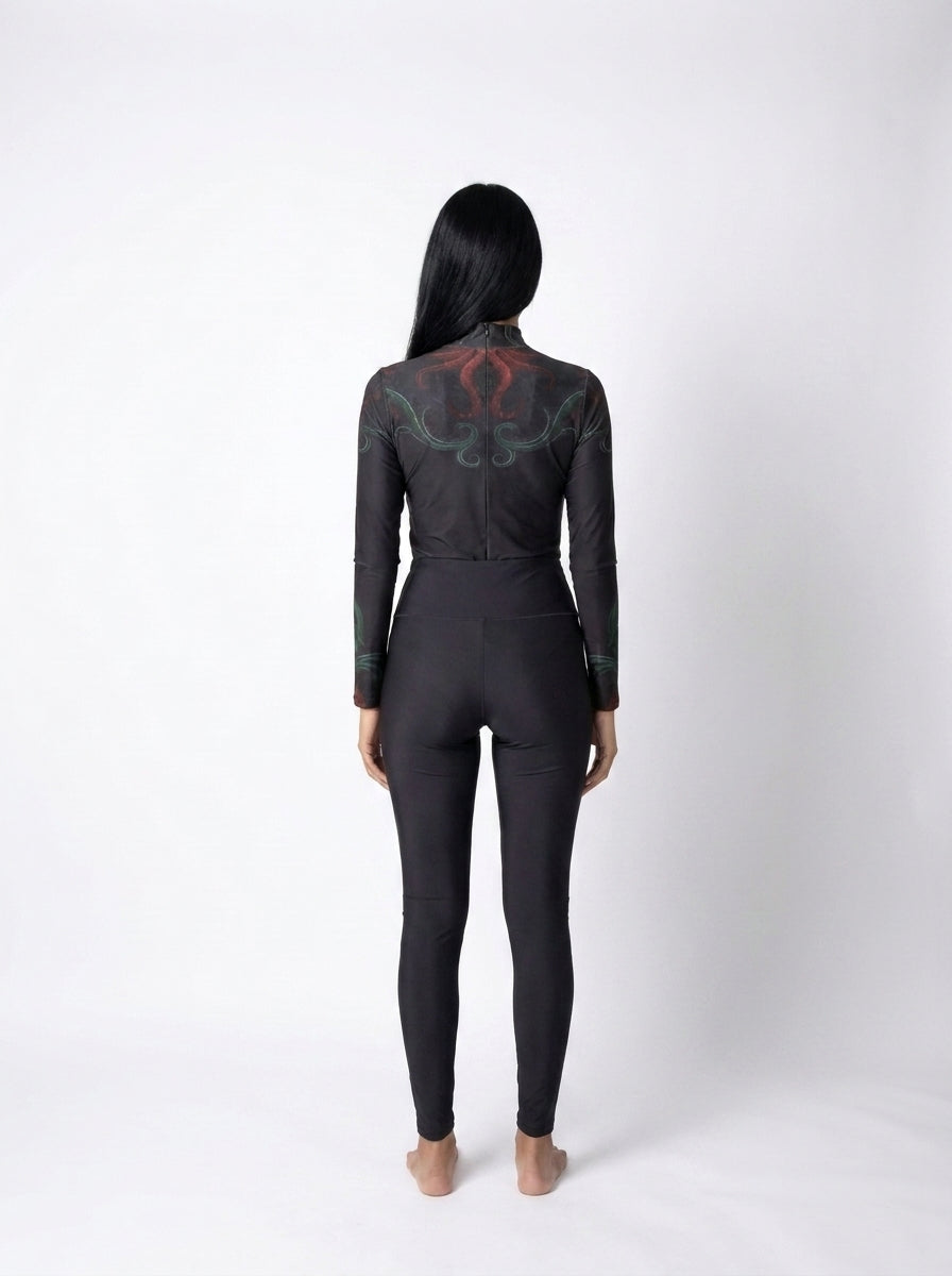 A woman viewed from the back wearing a black long-sleeve one-piece jumpsuit with a red and green dragon graphic across the upper back, standing against a light gray studio background.