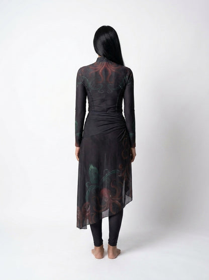A woman viewed from behind wearing a long-sleeve black sheer wrap dress with a red and green abstract print, paired with black leggings, against a plain white studio background.