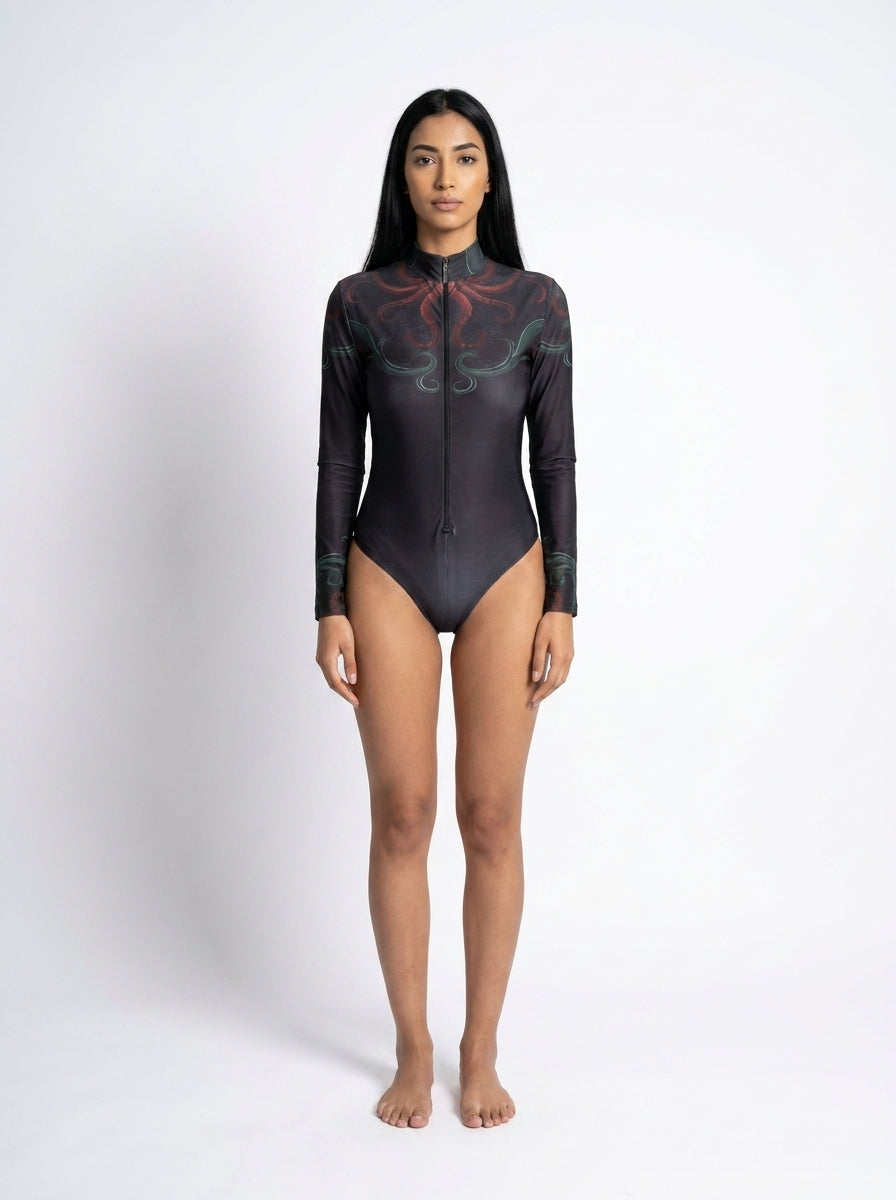 A woman stands facing forward in a studio wearing a long-sleeve black one-piece swimsuit with a subtle tonal graphic pattern and a high collar.