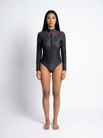 A woman stands facing forward in a studio wearing a long-sleeve black one-piece swimsuit with a subtle tonal graphic pattern and a high collar.