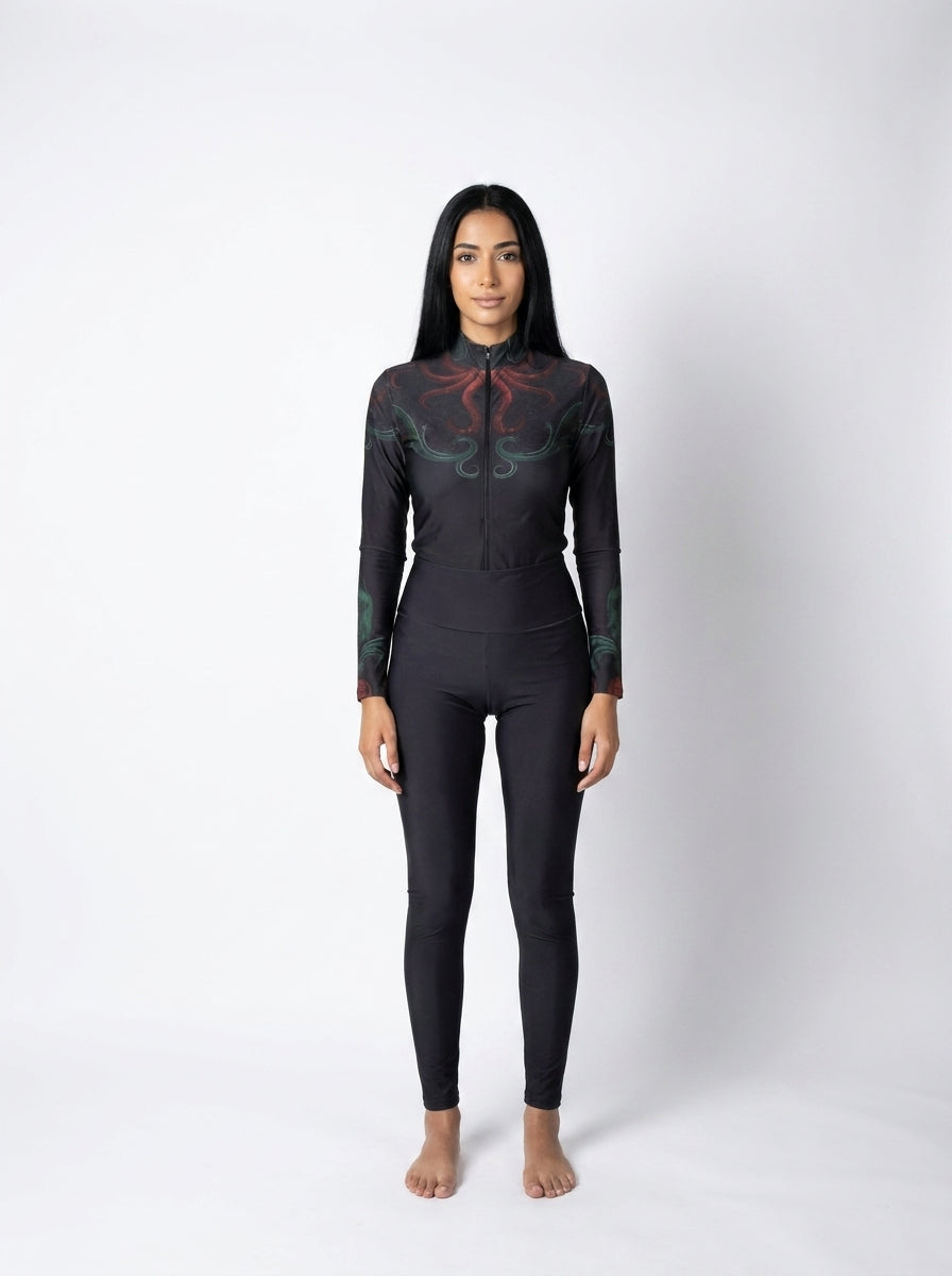 A woman stands barefoot against a light gray studio background wearing a black full-body wetsuit with a high collar and long sleeves, featuring subtle red and green abstract graphic details on the upper chest and shoulders.