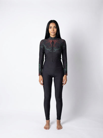 A woman stands barefoot against a light gray studio background wearing a black full-body wetsuit with a high collar and long sleeves, featuring subtle red and green abstract graphic details on the upper chest and shoulders.
