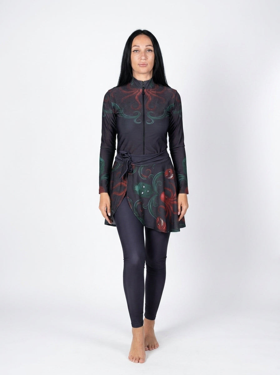 A woman modeling a long-sleeve swim dress with a high neck and a matching full-length swimsuit underneath, featuring a dark base with red and green abstract swirl patterns, against a plain white studio background.