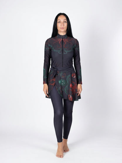 A woman modeling a long-sleeve swim dress with a high neck and a matching full-length swimsuit underneath, featuring a dark base with red and green abstract swirl patterns, against a plain white studio background.