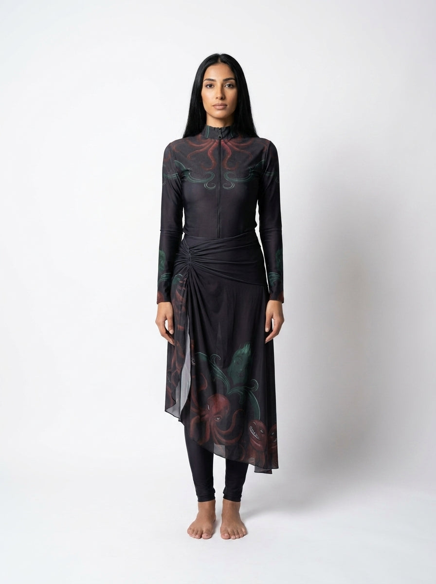 A woman stands against a light gray studio background wearing a long-sleeve black dress with a high mock neck, side tie detail, and a dark floral print in red and green. She is also wearing black leggings and is barefoot.