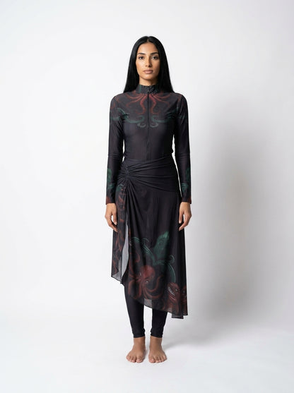 A woman stands against a light gray studio background wearing a long-sleeve black dress with a high mock neck, side tie detail, and a dark floral print in red and green. She is also wearing black leggings and is barefoot.