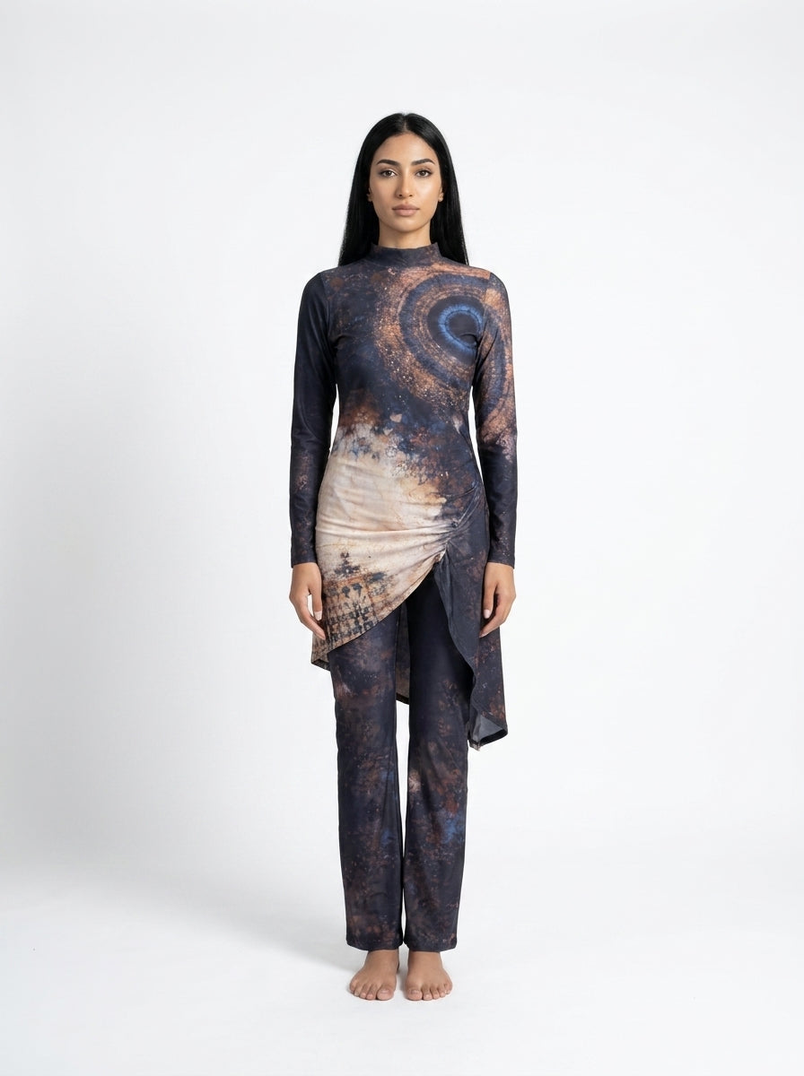 A woman stands barefoot against a light gray studio background wearing a long-sleeve, high-low asymmetrical tunic top and matching pants set. The fabric has an abstract cosmic print in dark navy, charcoal, and warm beige-brown tones.