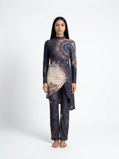 A woman stands barefoot against a light gray studio background wearing a long-sleeve, high-low asymmetrical tunic top and matching pants set. The fabric has an abstract cosmic print in dark navy, charcoal, and warm beige-brown tones.