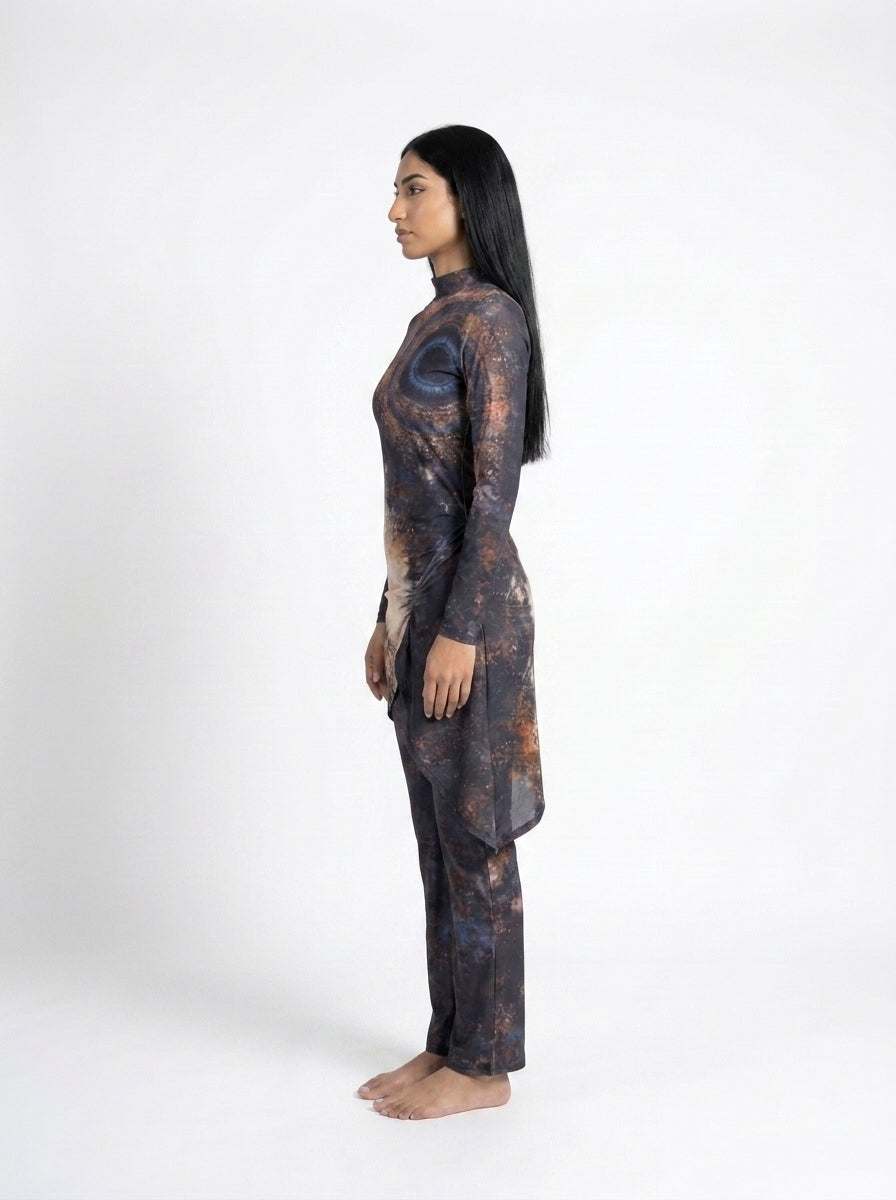 A woman stands in profile against a plain white studio background, wearing a long-sleeve tunic and matching pants with an abstract dark print in black, brown, and muted blue.