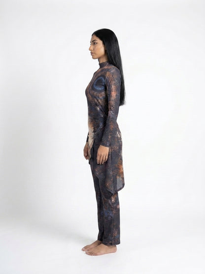 A woman stands in profile against a plain white studio background, wearing a long-sleeve tunic and matching pants with an abstract dark print in black, brown, and muted blue.