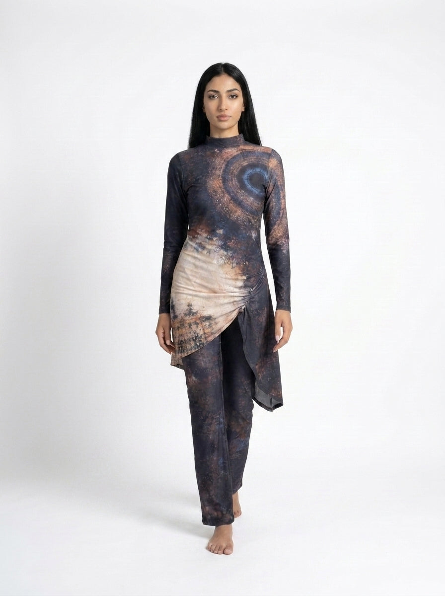 A woman modeling a long-sleeve, high-waisted, asymmetrical wrap-style tunic and matching pants set in a dark abstract print with brown, beige, and blue tones, shot against a plain white studio background.