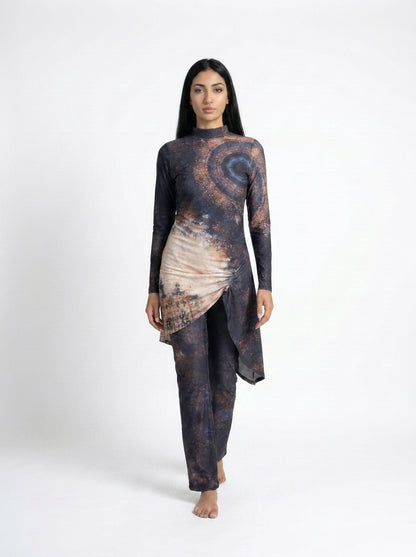 A woman modeling a long-sleeve, high-waisted, asymmetrical wrap-style tunic and matching pants set in a dark abstract print with brown, beige, and blue tones, shot against a plain white studio background.