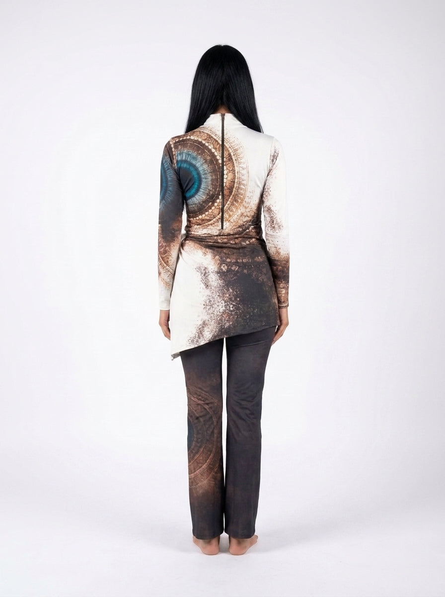 A woman viewed from the back wearing a long-sleeve asymmetrical top with a brown and blue abstract spiral print and dark charcoal pants, standing against a plain white studio background.