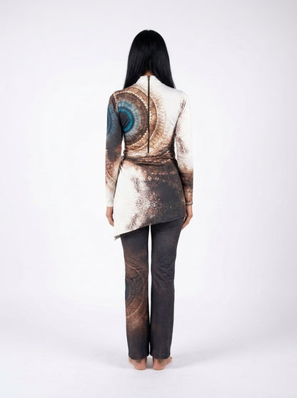 A woman viewed from the back wearing a long-sleeve asymmetrical top with a brown and blue abstract spiral print and dark charcoal pants, standing against a plain white studio background.