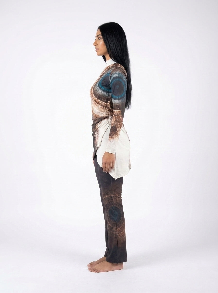 A woman stands in profile against a plain white studio background, wearing a loose long-sleeve top with a brown, beige, and blue abstract print and dark brown pants with a matching abstract print.