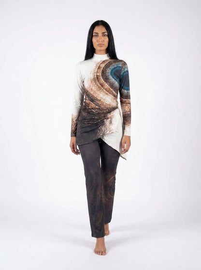 A woman modeling a long-sleeve asymmetrical top with a brown, beige, and blue abstract swirl print, paired with dark charcoal pants, against a plain white studio background.