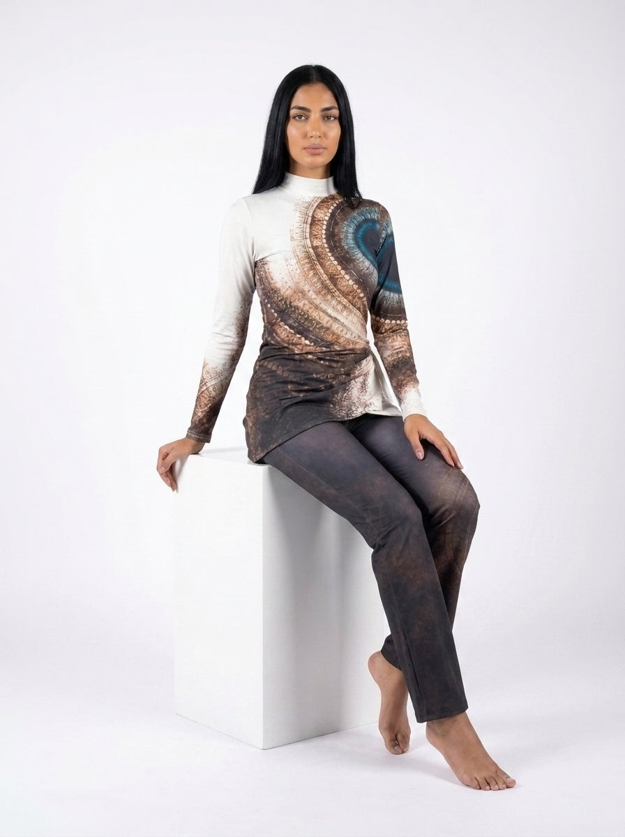 A woman sits on a white cube in a studio setting, wearing a long-sleeve mock-neck top with a brown and blue spiral fractal pattern and dark gray patterned leggings.