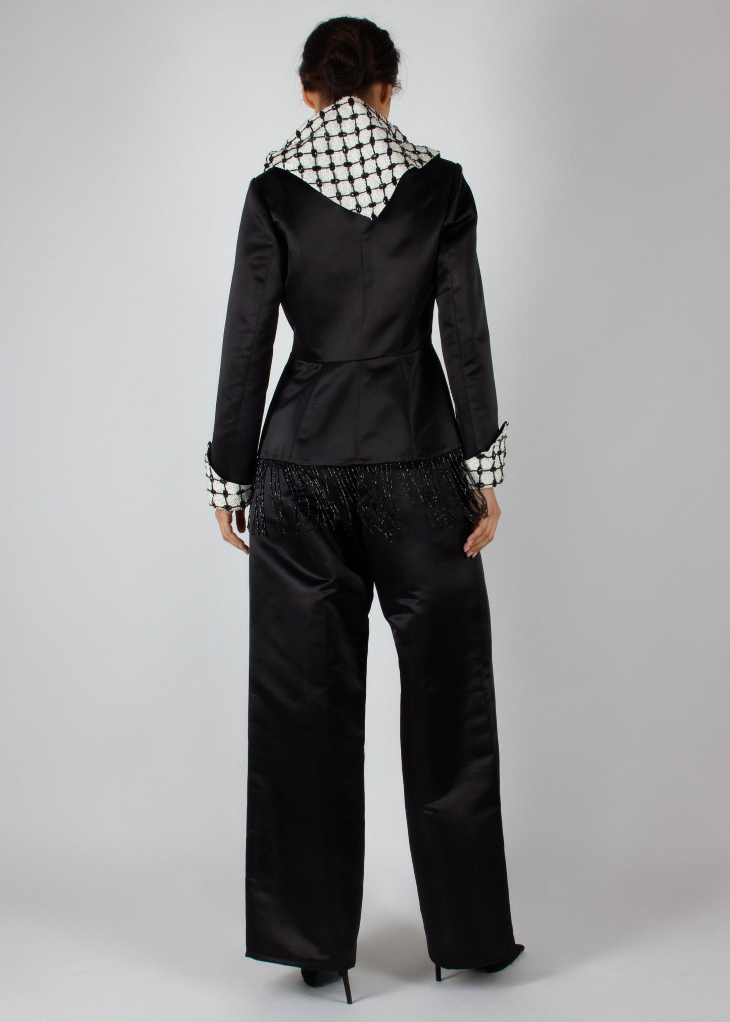 Elegant black satin pantsuit from the HORRA collection by MNour Design, featuring a statement keffiyeh-inspired lapel and cuff details. The tailored blazer is adorned with intricate beaded fringe, adding a touch of modern sophistication. Discover more at www.mnourdesign.com