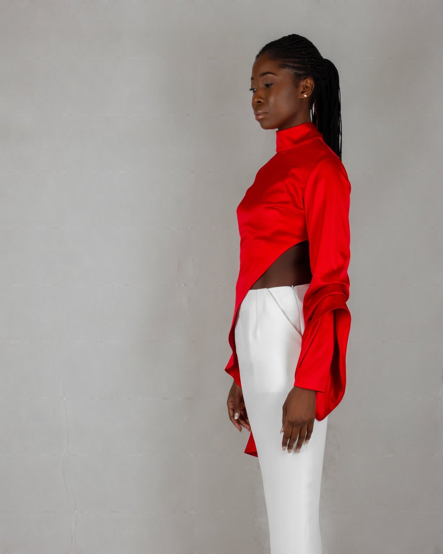 A model showcases a striking outfit from the "HAWAA" collection, specially designed for Valentine's Day. The ensemble features a bold red, high-neck blouse with long sleeves and an asymmetrical design that drapes elegantly over one side. It is paired with sleek white wide-leg pants. The model stands barefoot against a minimalist gray textured background, highlighting the vibrant contrast between the red top and white pants.