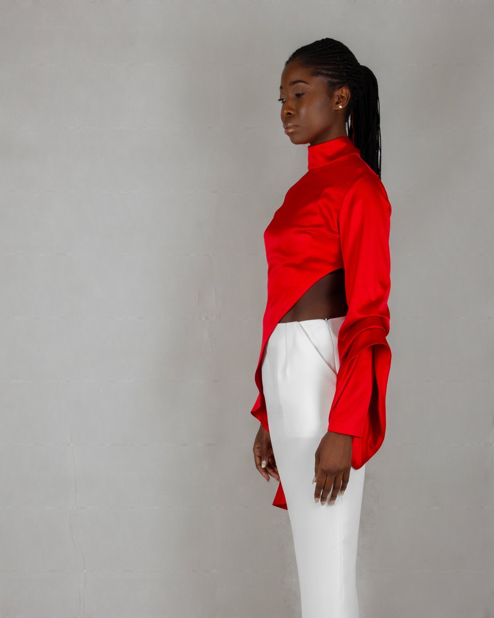 A model showcases a striking outfit from the "HAWAA" collection, specially designed for Valentine's Day. The ensemble features a bold red, high-neck blouse with long sleeves and an asymmetrical design that drapes elegantly over one side. It is paired with sleek white wide-leg pants. The model stands barefoot against a minimalist gray textured background, highlighting the vibrant contrast between the red top and white pants.