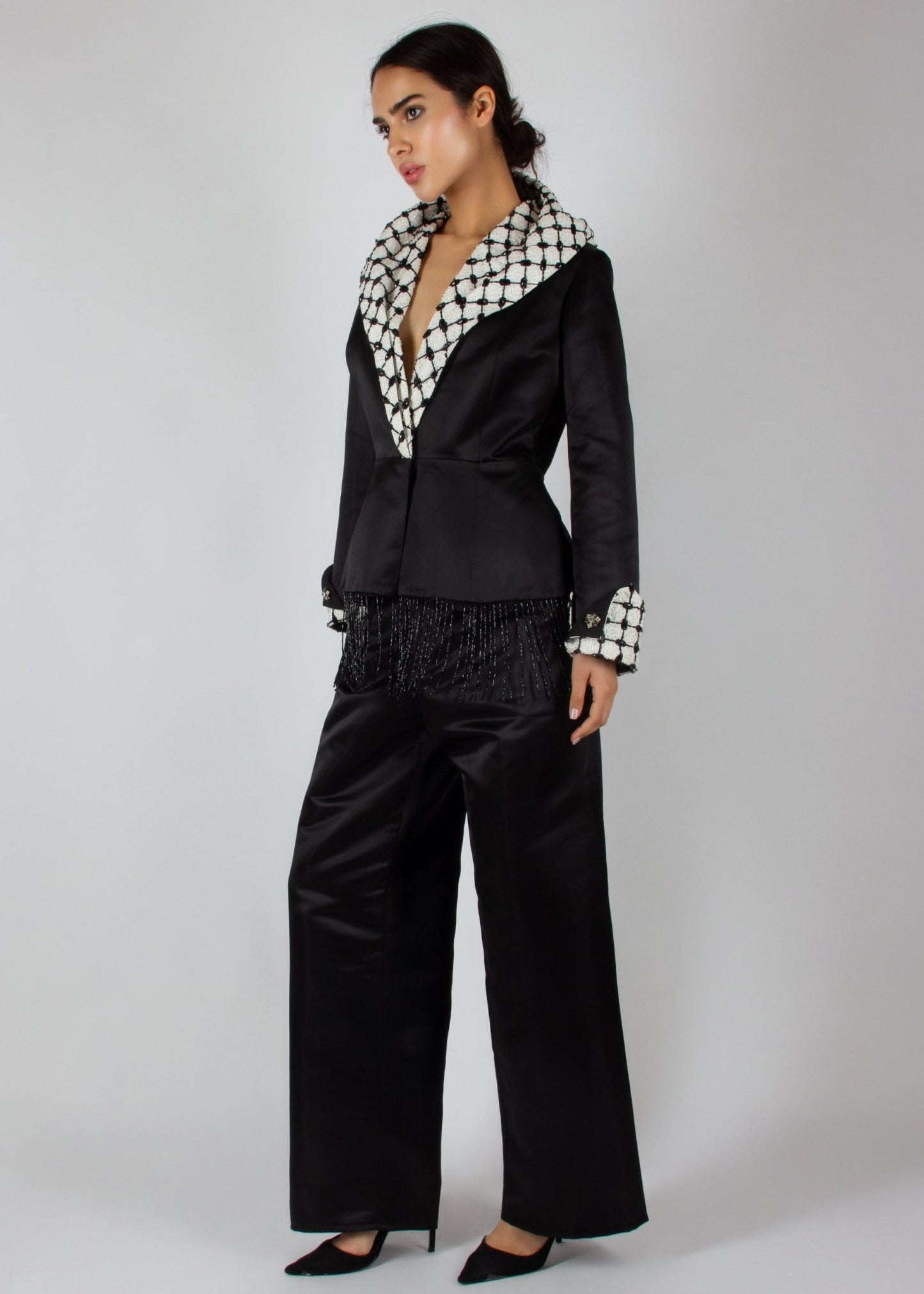 Elegant black satin pantsuit from the HORRA collection by MNour Design, featuring a statement keffiyeh-inspired lapel and cuff details. The tailored blazer is adorned with intricate beaded fringe, adding a touch of modern sophistication. Discover more at www.mnourdesign.com