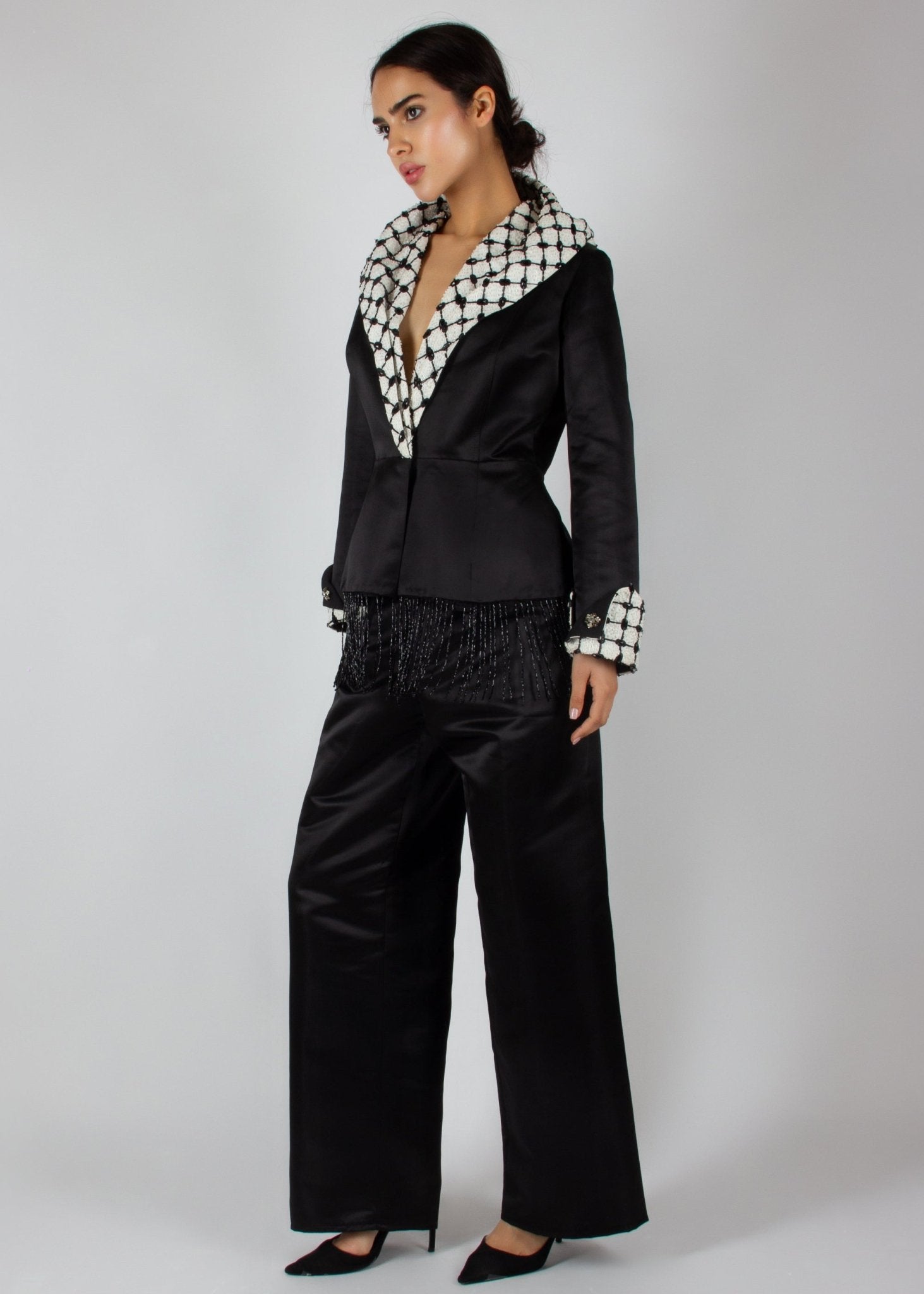 Elegant black satin pantsuit from the HORRA collection by MNour Design, featuring a statement keffiyeh-inspired lapel and cuff details. The tailored blazer is adorned with intricate beaded fringe, adding a touch of modern sophistication. Discover more at www.mnourdesign.com
