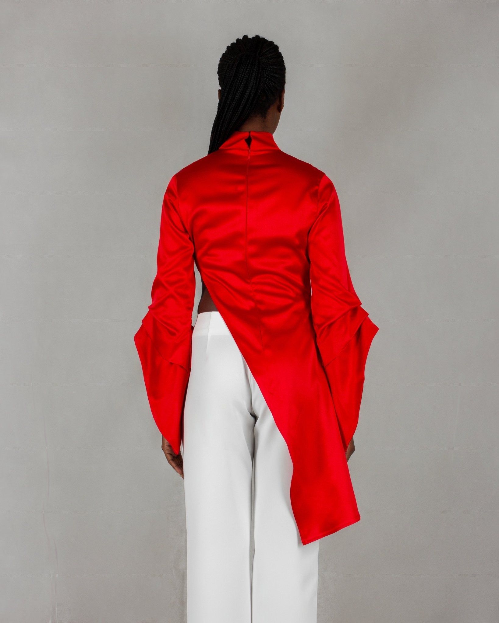 A model showcases a striking outfit from the "HAWAA" collection, specially designed for Valentine's Day. The ensemble features a bold red, high-neck blouse with long sleeves and an asymmetrical design that drapes elegantly over one side. It is paired with sleek white wide-leg pants. The model stands barefoot against a minimalist gray textured background, highlighting the vibrant contrast between the red top and white pants.