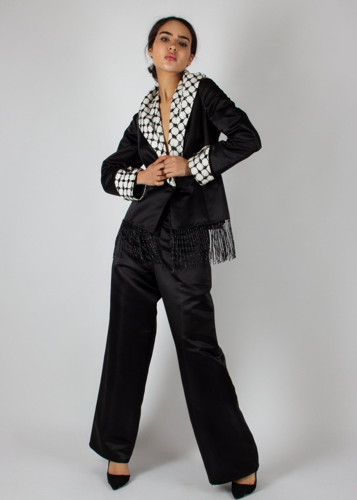 Elegant black satin pantsuit from the HORRA collection by MNour Design, featuring a statement keffiyeh-inspired lapel and cuff details. The tailored blazer is adorned with intricate beaded fringe, adding a touch of modern sophistication. Discover more at www.mnourdesign.com