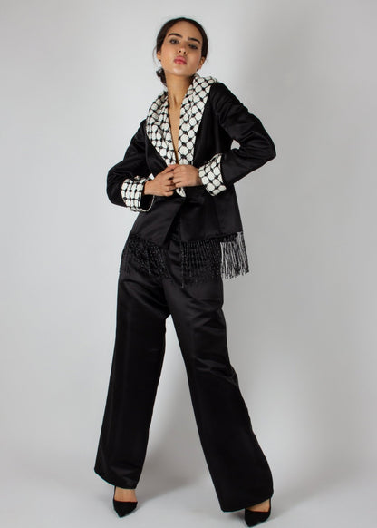 Elegant black satin pantsuit from the HORRA collection by MNour Design, featuring a statement keffiyeh-inspired lapel and cuff details. The tailored blazer is adorned with intricate beaded fringe, adding a touch of modern sophistication. Discover more at www.mnourdesign.com