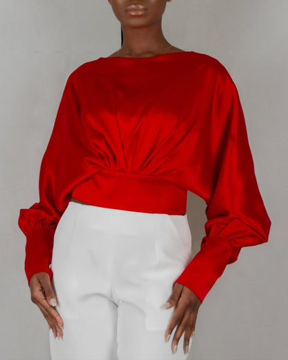 A model wears a bold red, satin-like top from the "HAWAA" collection, specially designed for Valentine's Day. The top features an asymmetrical design with puffed sleeves and a draped neckline that creates a cut-out detail on one side. It is cinched at the waist with a matching belt and paired with black pants. The model stands against a simple gray textured background, emphasizing the vibrant color and elegant silhouette of the outfit.