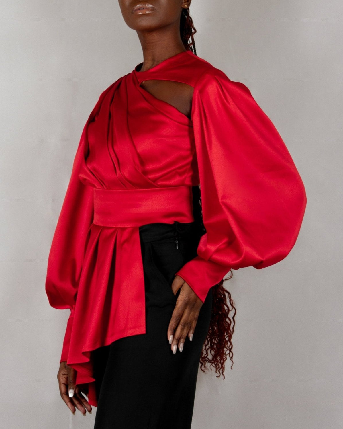 A model wears a bold red, satin-like top from the "HAWAA" collection, specially designed for Valentine's Day. The top features an asymmetrical design with puffed sleeves and a draped neckline that creates a cut-out detail on one side. It is cinched at the waist with a matching belt and paired with black pants. The model stands against a simple gray textured background, emphasizing the vibrant color and elegant silhouette of the outfit.
