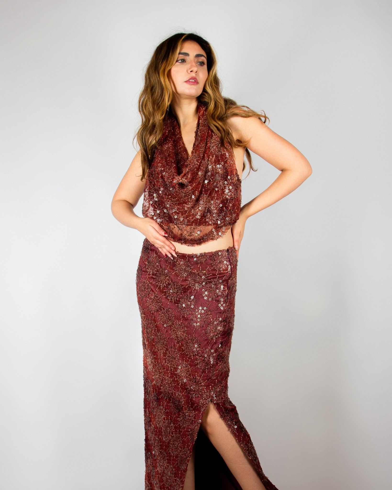 Elegant two-piece evening ensemble by MNour Design, featuring a shimmering burgundy sequined skirt with a high slit and a draped cowl-neck crop top. The intricate beadwork and rich fabric exude sophistication and modern glamour. Shop this exclusive design at www.mnourdesign.com
