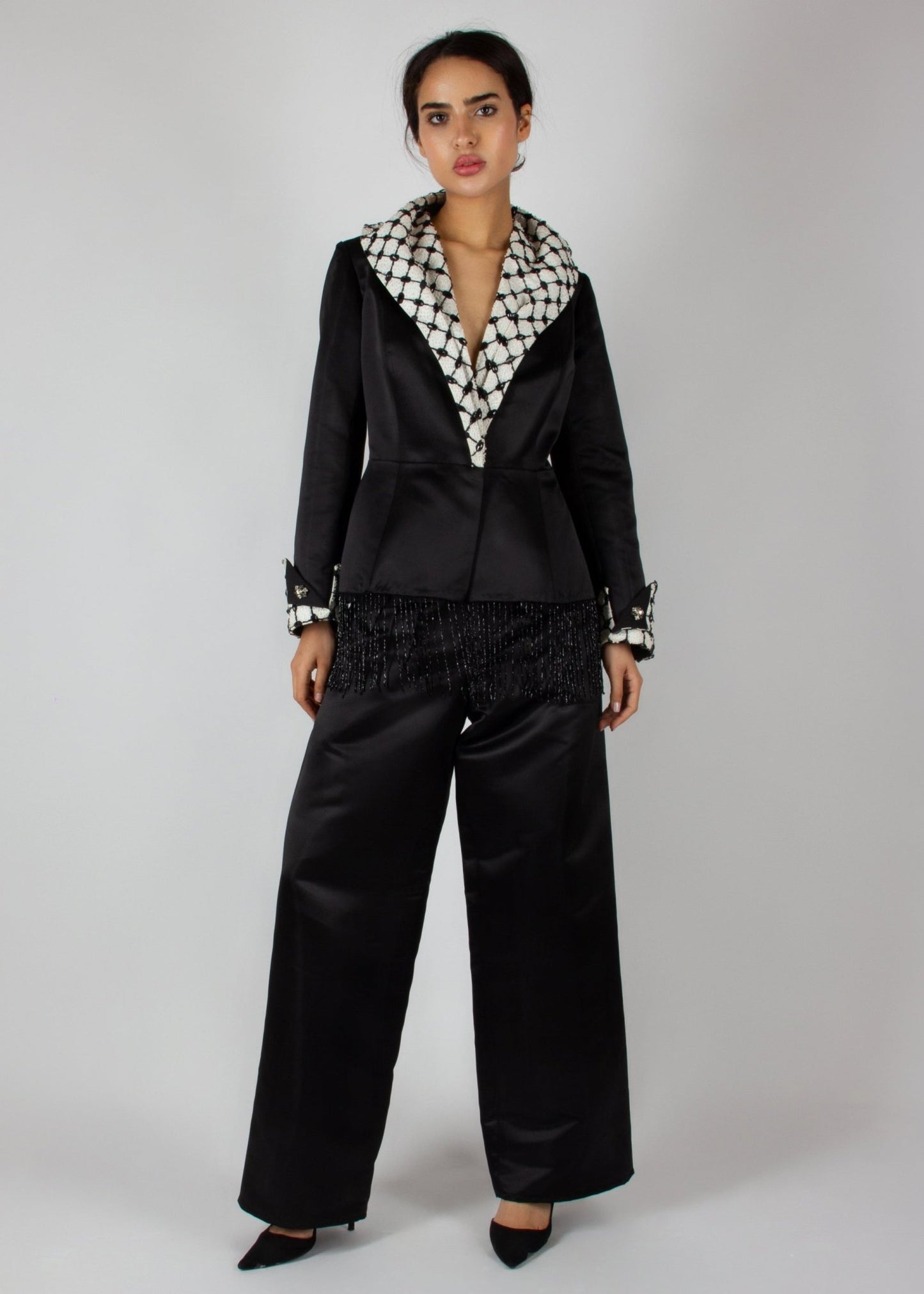 Elegant black satin pantsuit from the HORRA collection by MNour Design, featuring a statement keffiyeh-inspired lapel and cuff details. The tailored blazer is adorned with intricate beaded fringe, adding a touch of modern sophistication. Discover more at www.mnourdesign.com