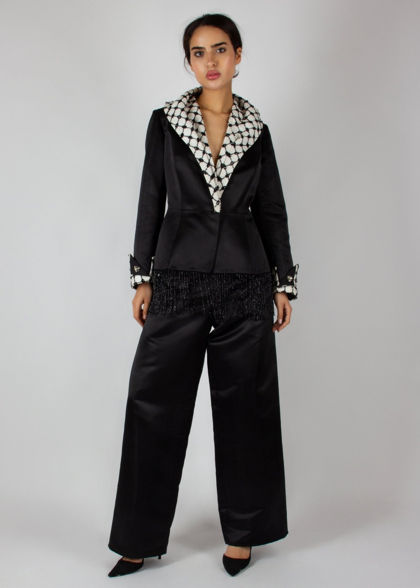 Elegant black satin pantsuit from the HORRA collection by MNour Design, featuring a statement keffiyeh-inspired lapel and cuff details. The tailored blazer is adorned with intricate beaded fringe, adding a touch of modern sophistication. Discover more at www.mnourdesign.com