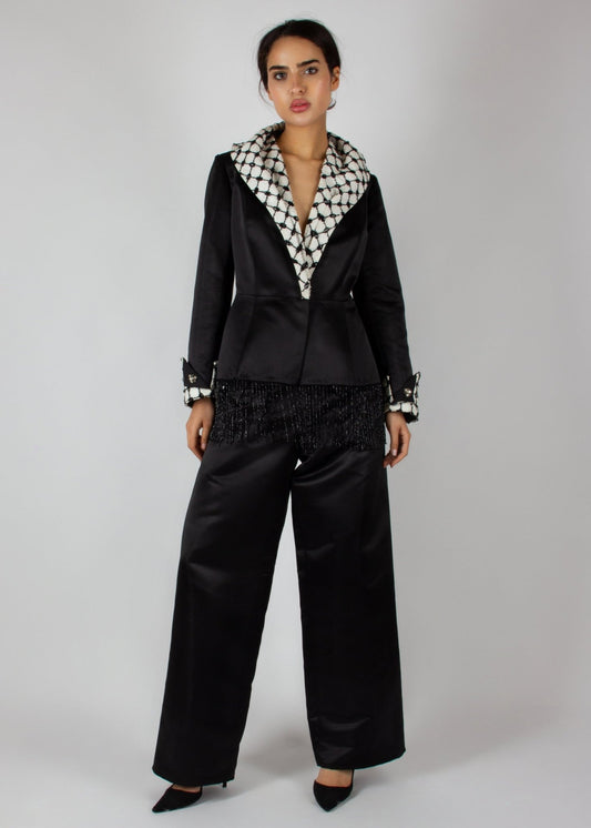 Elegant black satin pantsuit from the HORRA collection by MNour Design, featuring a statement keffiyeh-inspired lapel and cuff details. The tailored blazer is adorned with intricate beaded fringe, adding a touch of modern sophistication. Discover more at www.mnourdesign.com