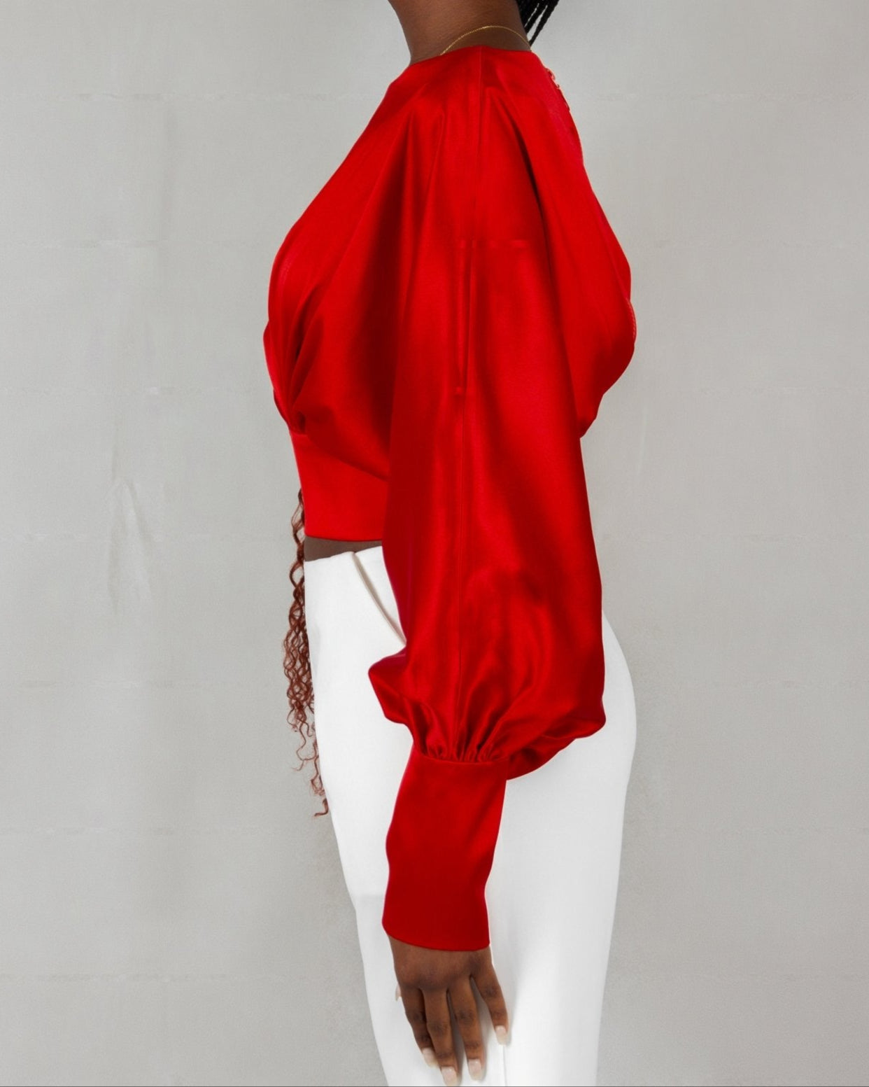 A model wears a bold red, satin-like top from the "HAWAA" collection, specially designed for Valentine's Day. The top features an asymmetrical design with puffed sleeves and a draped neckline that creates a cut-out detail on one side. It is cinched at the waist with a matching belt and paired with black pants. The model stands against a simple gray textured background, emphasizing the vibrant color and elegant silhouette of the outfit.
