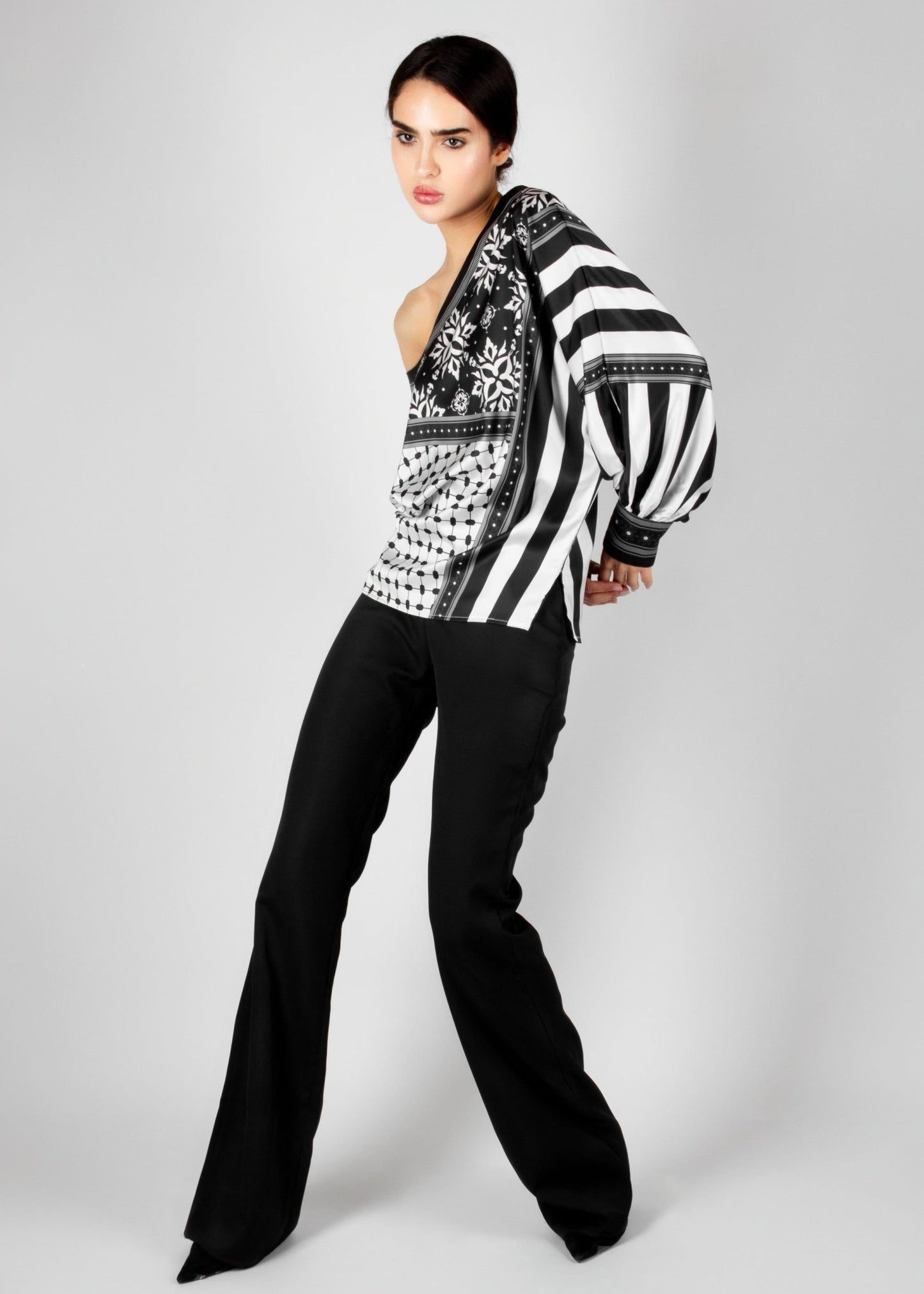 Bold off-shoulder blouse from the HORRA collection by MNour Design, featuring a striking keffiyeh-inspired pattern with intricate geometric details. Paired with sleek black trousers for a modern, sophisticated look. Explore more at www.mnourdesign.com