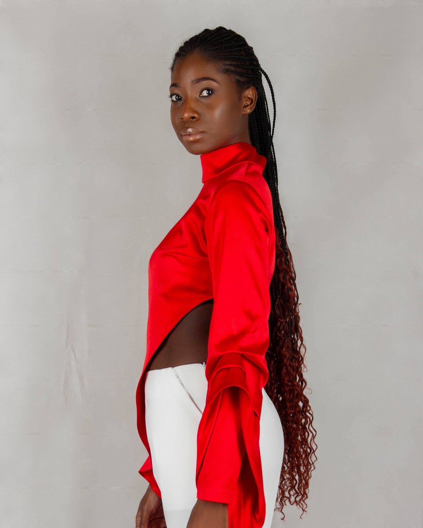 A model showcases a striking outfit from the "HAWAA" collection, specially designed for Valentine's Day. The ensemble features a bold red, high-neck blouse with long sleeves and an asymmetrical design that drapes elegantly over one side. It is paired with sleek white wide-leg pants. The model stands barefoot against a minimalist gray textured background, highlighting the vibrant contrast between the red top and white pants.