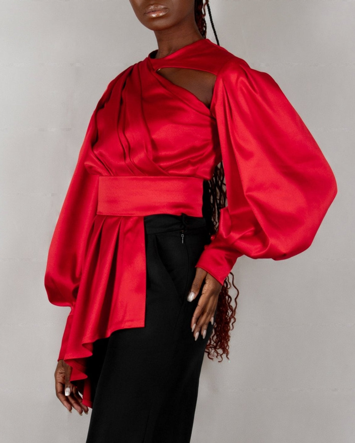 A model wears a bold red, satin-like top from the "HAWAA" collection, specially designed for Valentine's Day. The top features an asymmetrical design with puffed sleeves and a draped neckline that creates a cut-out detail on one side. It is cinched at the waist with a matching belt and paired with black pants. The model stands against a simple gray textured background, emphasizing the vibrant color and elegant silhouette of the outfit.