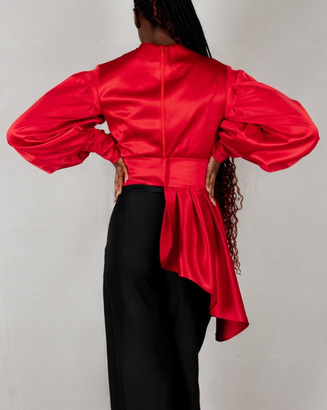 A model wears a bold red, satin-like top from the "HAWAA" collection, specially designed for Valentine's Day. The top features an asymmetrical design with puffed sleeves and a draped neckline that creates a cut-out detail on one side. It is cinched at the waist with a matching belt and paired with black pants. The model stands against a simple gray textured background, emphasizing the vibrant color and elegant silhouette of the outfit.