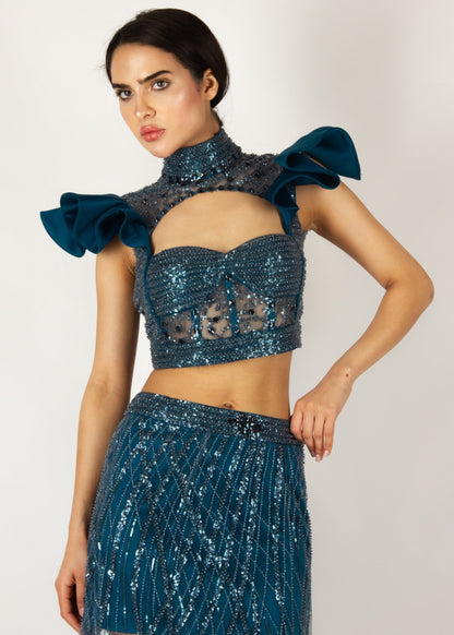 Elegant two-piece couture set by MNour Design, featuring a shimmering blue embellished crop top with dramatic ruffled shoulders and a high-neck cut, paired with a matching sheer beaded skirt for a bold and sophisticated look. Explore this stunning design at www.mnourdesign.com.