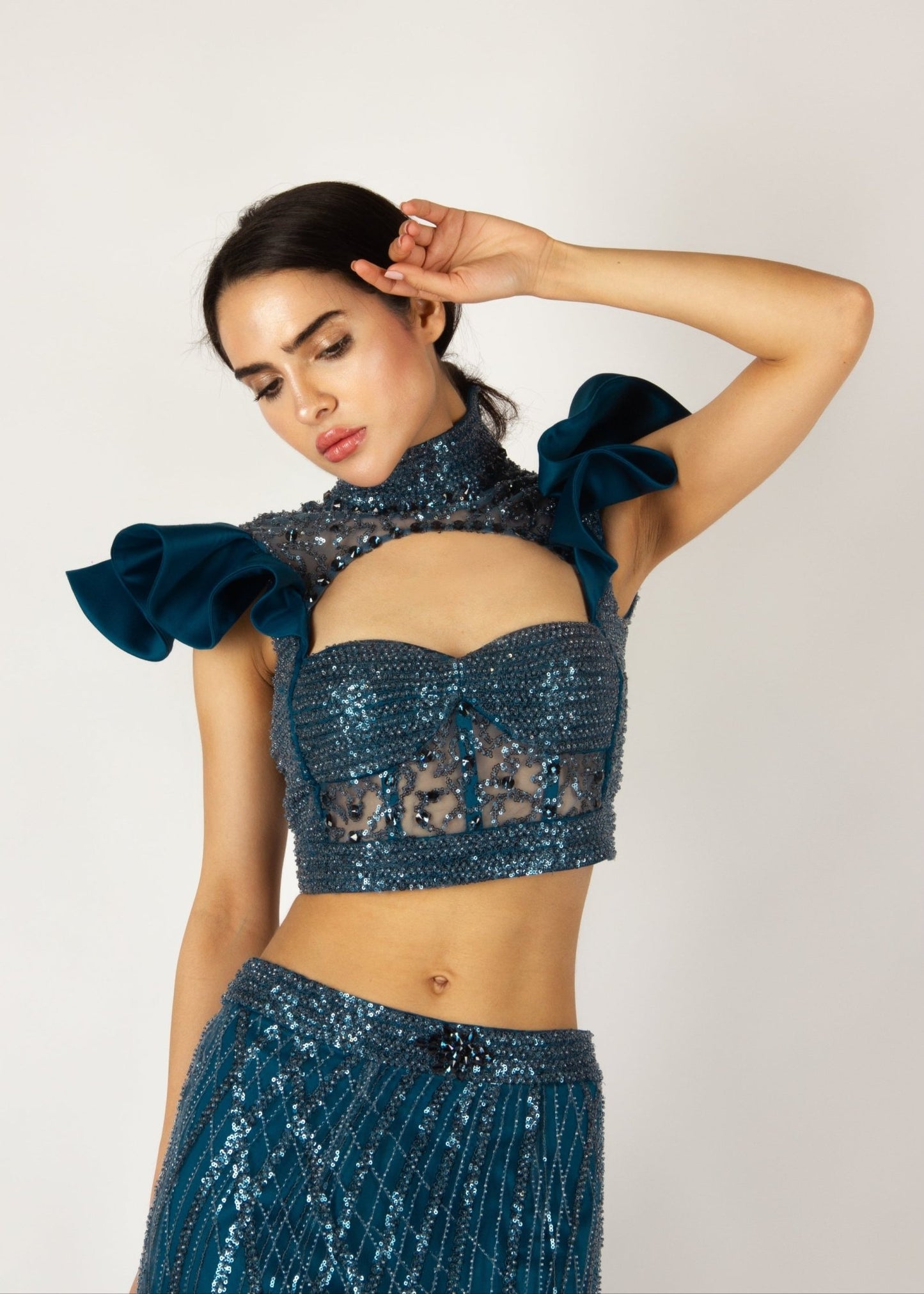 Elegant two-piece couture set by MNour Design, featuring a shimmering blue embellished crop top with dramatic ruffled shoulders and a high-neck cut, paired with a matching sheer beaded skirt for a bold and sophisticated look. Explore this stunning design at www.mnourdesign.com.