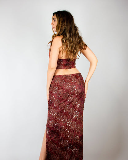 Elegant two-piece evening ensemble by MNour Design, featuring a shimmering burgundy sequined skirt with a high slit and a draped cowl-neck crop top. The intricate beadwork and rich fabric exude sophistication and modern glamour. Shop this exclusive design at www.mnourdesign.com