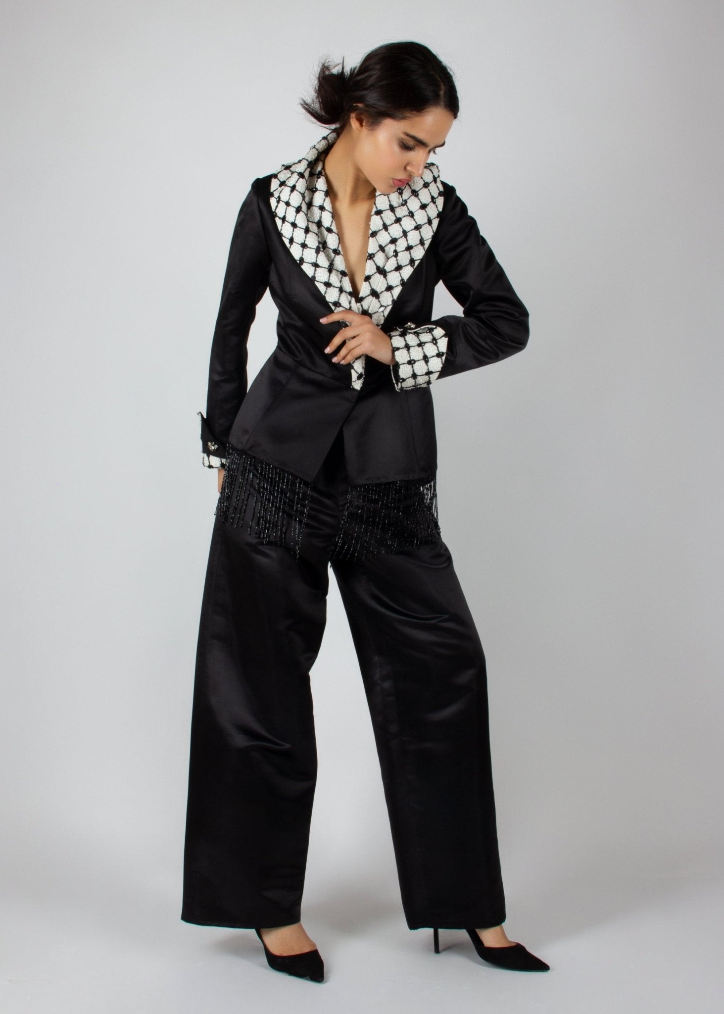 Elegant black satin pantsuit from the HORRA collection by MNour Design, featuring a statement keffiyeh-inspired lapel and cuff details. The tailored blazer is adorned with intricate beaded fringe, adding a touch of modern sophistication. Discover more at www.mnourdesign.com