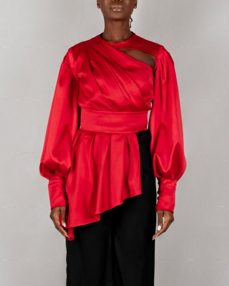 A model wears a bold red, satin-like top from the "HAWAA" collection, specially designed for Valentine's Day. The top features an asymmetrical design with puffed sleeves and a draped neckline that creates a cut-out detail on one side. It is cinched at the waist with a matching belt and paired with black pants. The model stands against a simple gray textured background, emphasizing the vibrant color and elegant silhouette of the outfit.