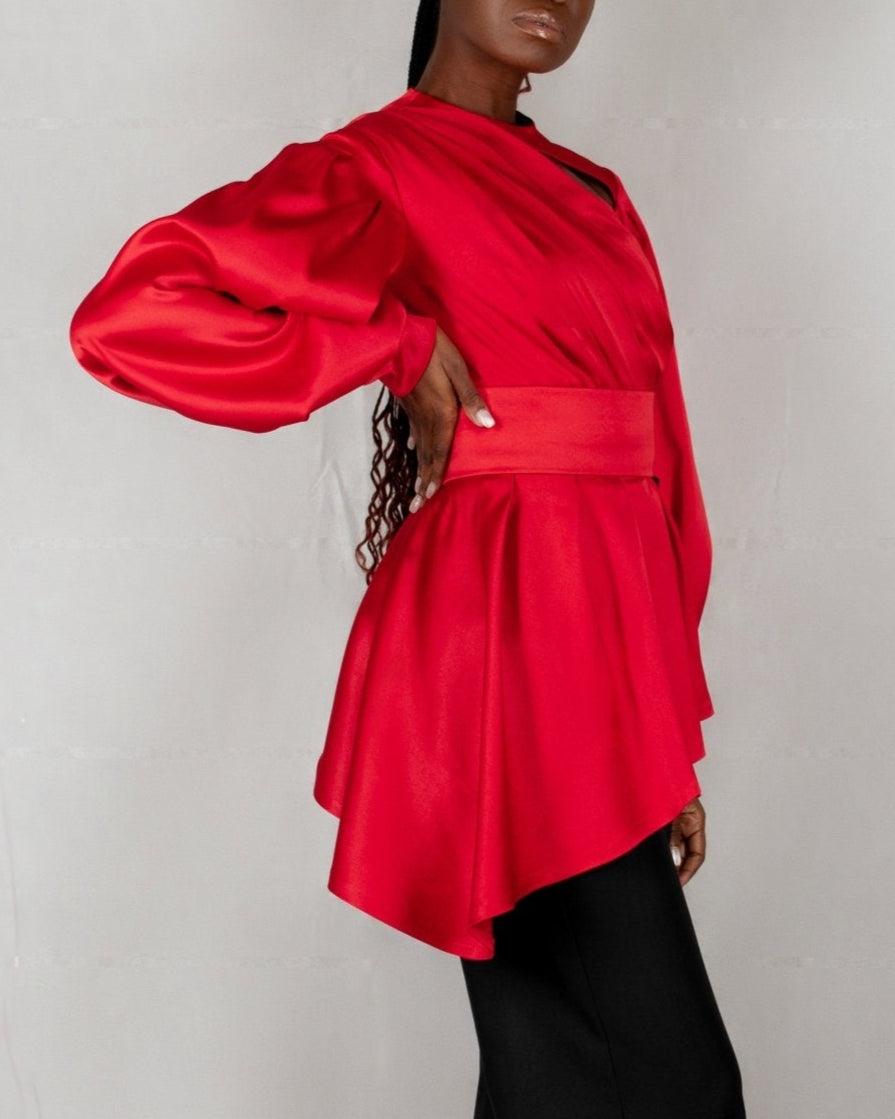A model wears a bold red, satin-like top from the "HAWAA" collection, specially designed for Valentine's Day. The top features an asymmetrical design with puffed sleeves and a draped neckline that creates a cut-out detail on one side. It is cinched at the waist with a matching belt and paired with black pants. The model stands against a simple gray textured background, emphasizing the vibrant color and elegant silhouette of the outfit.