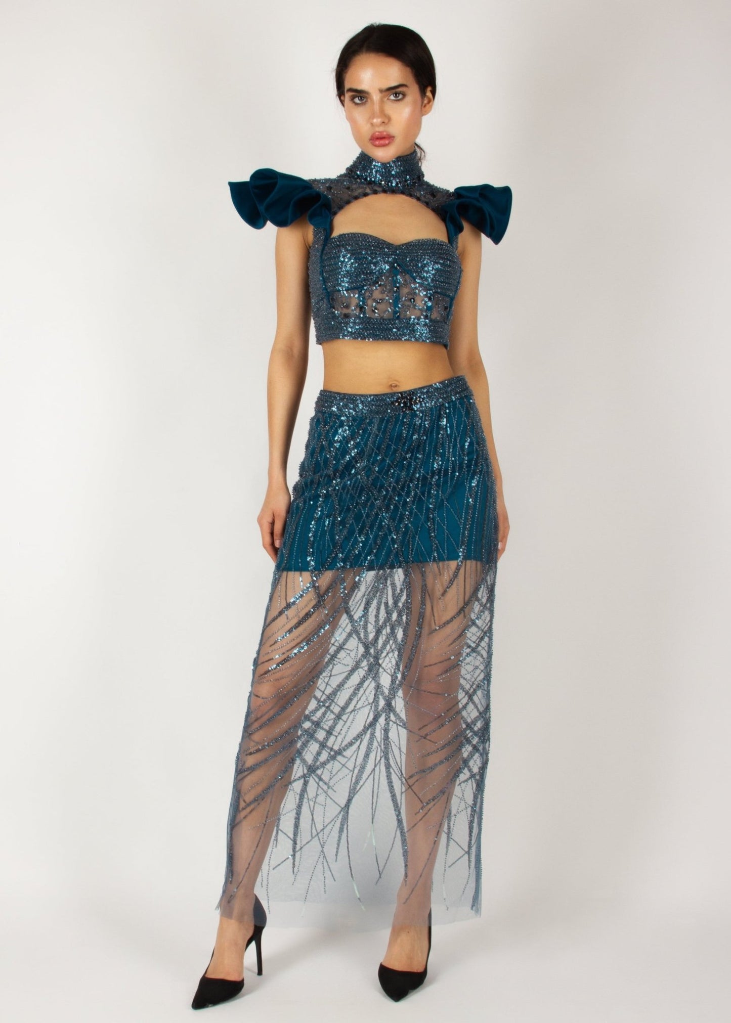 Elegant two-piece couture set by MNour Design, featuring a shimmering blue embellished crop top with dramatic ruffled shoulders and a high-neck cut, paired with a matching sheer beaded skirt for a bold and sophisticated look. Explore this stunning design at www.mnourdesign.com.